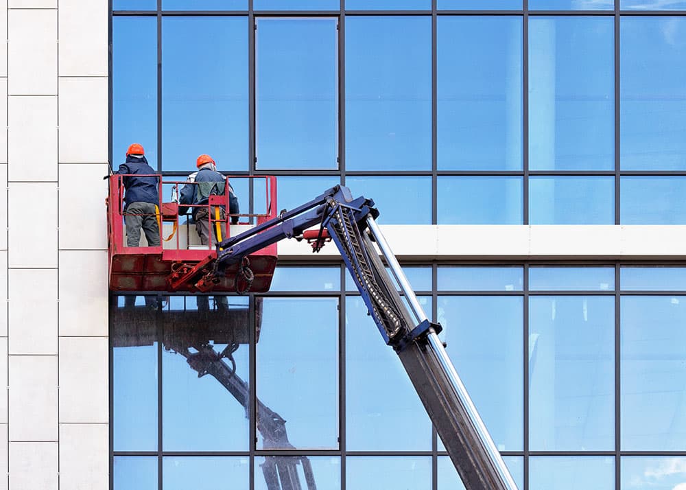 Professionelle Glasfassadenreinigung in Hamburg: Zwei Gebäudereiniger mit Sicherheitsausrüstung reinigen eine hoch gelegene Glasfront aus einem Hubsteiger heraus
