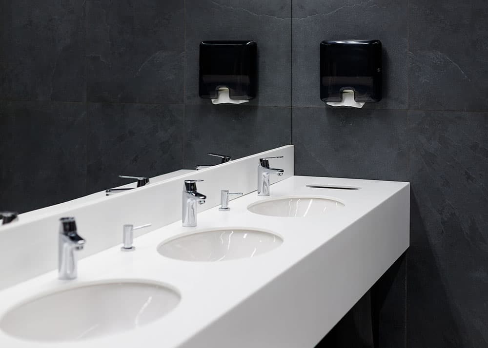 Hygienisch sauberer Waschbereich mit drei Waschbecken, Wandspiegel und Papierhandtuchspender – professionelle Sanitärreinigung für Gewerbeobjekte